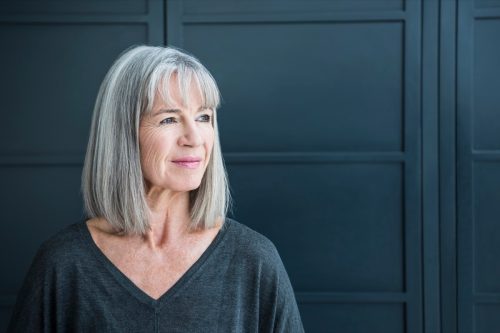 One senior woman with a grey bob in front of a dark grey background. Portrait of smart senior woman in her 60s, looking away from camera.