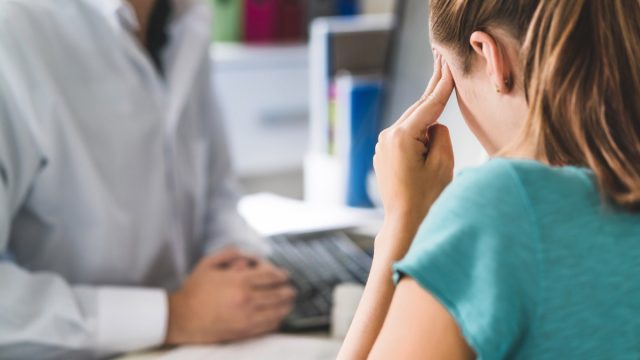 Woman suffering from bad headache or migraine. Appointment with doctor in office room. Sick and unwell lady with stress, trauma or burnout. Young patient in pain holding head with hands. Medical visit