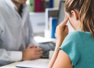 Woman suffering from bad headache or migraine. Appointment with doctor in office room. Sick and unwell lady with stress, trauma or burnout. Young patient in pain holding head with hands. Medical visit