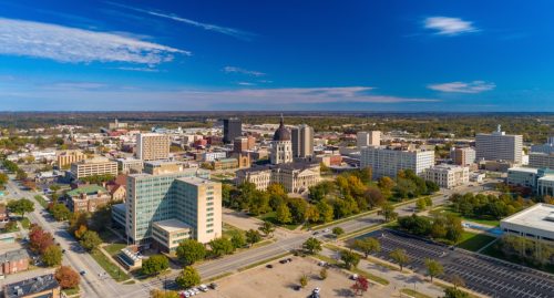 Downtown Topeka aerial skyline view with the Kansas State Capitol building in the center, and a blue sky with clouds in the background.