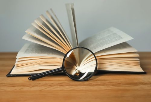 Magnifying glass and open books with turning pages on wood table.