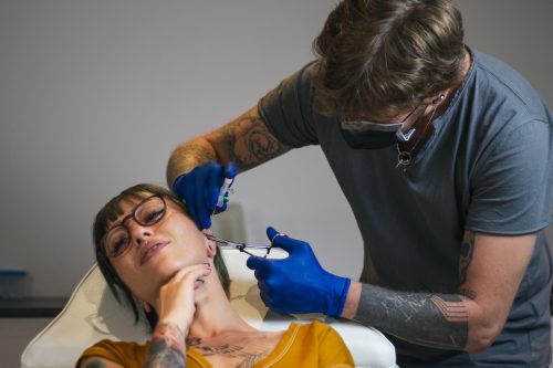 Young Woman getting her ear pierced. Man showing a process of piercing with steril medical equipment and latex gloves.