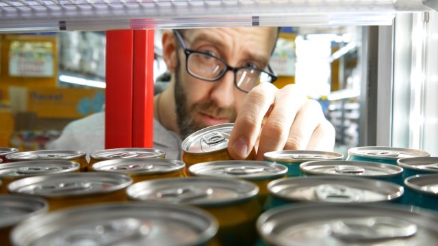 Close-up of the hand of a bearded man with glasses taking one can of soda or energy drink in a store fridge