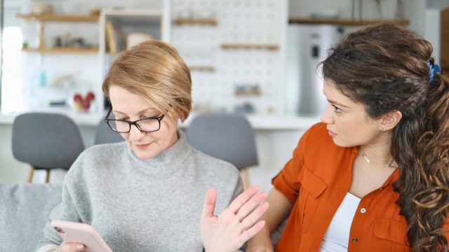 Shot of a senior mother looking at the phone while upset daughter looks at her on the couch.