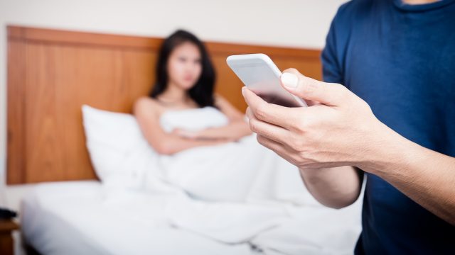 Cheating concept with unhappy couple – woman in bed while husband or boyfriend is texting