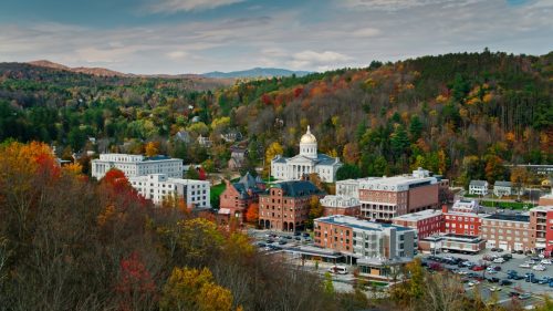 Aerial shot of the state capitol building in Montpelier, Vermont in Fall, flying over downtown buildings with a mountainside covered in colorful autumnal trees behind.