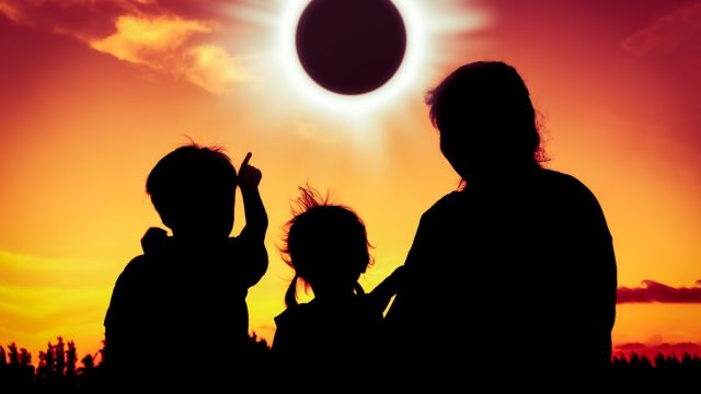 Silhouette back view of family sitting and relaxing together. Boy point to solar eclipse on gold sky background. Happy family spending time together. Outdoor.