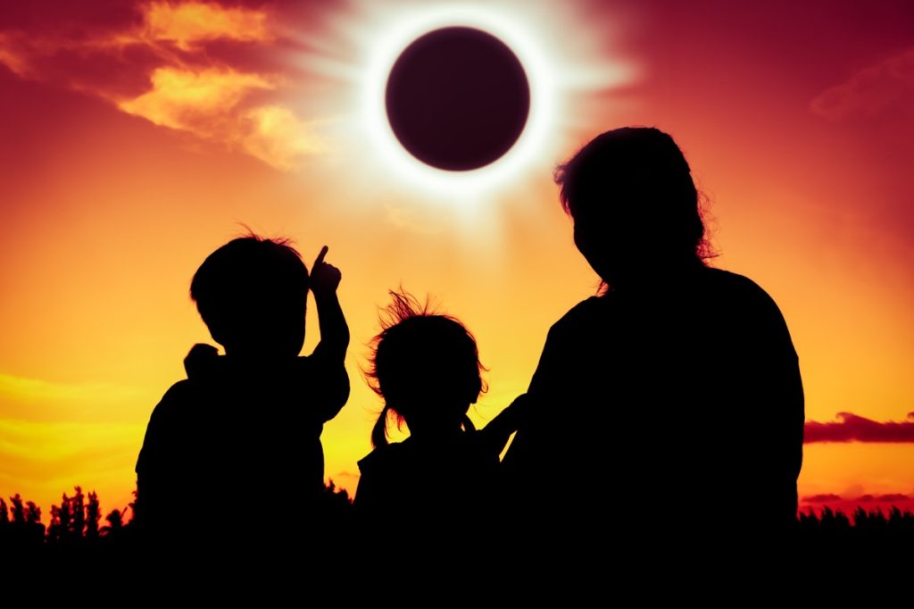 Silhouette back view of family sitting and relaxing together. Boy point to solar eclipse on gold sky background. Happy family spending time together. Outdoor.
