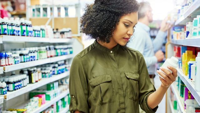 Pharmacy, health shop and woman with medicine for reading label, check product and choice in retail store. African female person looking at drugstore pills for ingredients, information and shopping