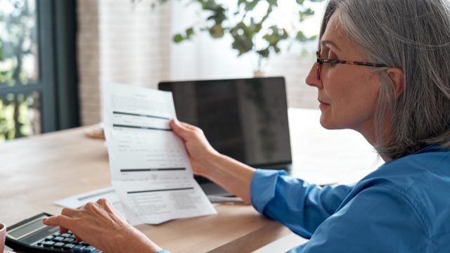Senior woman holding a paper bill using a calculator at her desk