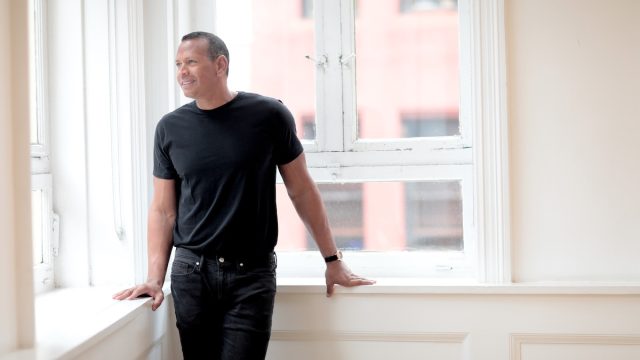 Alex Rodriguez wearing black jeans and a black t-shirt standing against white walls and large, city windows
