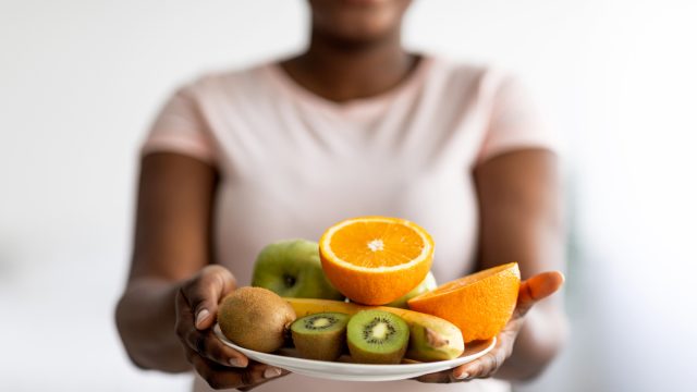 Blurred woman wearing a pale pink t-shirt holding out a plate of fruit, including oranges, kiwi, and banana.