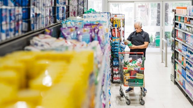 Senior man with shopping cart looking at grocery products in supermarket aisle. Mature male customer looking at groceries in store.