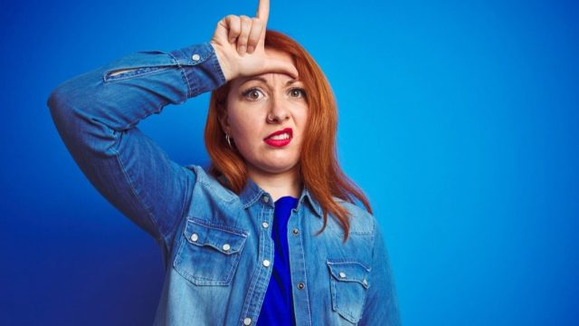 woman doing the "loser" sign on her forehead against a blue background