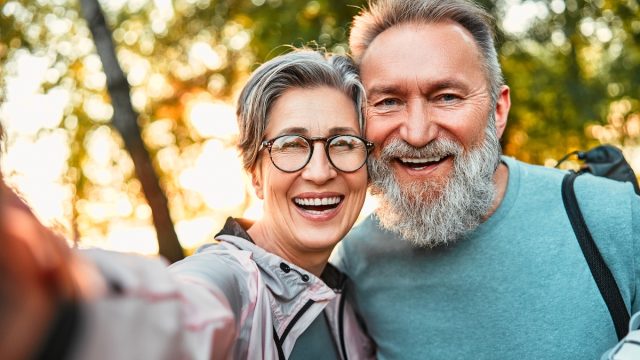 A happy, mature couple both with gray hair take a smiling selfie while standing outside among trees