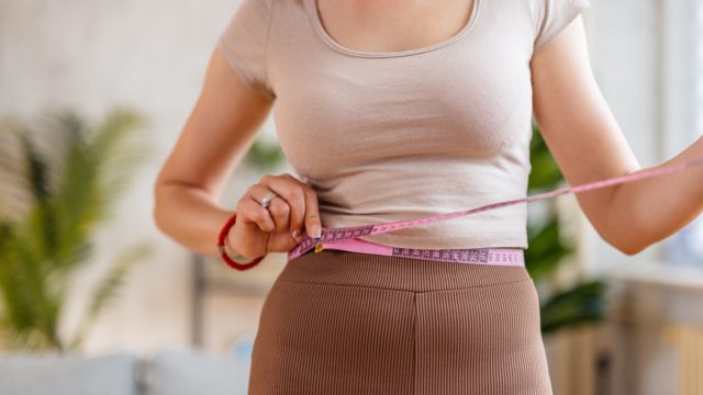 Beautiful young woman measuring her waist with a measuring tape in the living room at home.