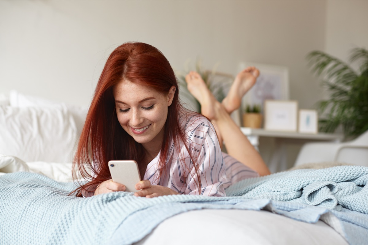 woman with red hair laying on her stomach in bed, using her phone