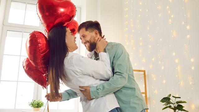 A happy, romantic couple hugging in their living room; the man is holding red heart-shaped balloons