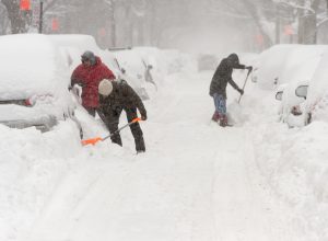 Three people shoveling out cars on a street during a heavy snowstorm