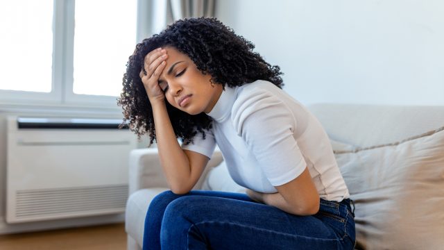 A woman sitting on the couch and holding her stomach and head in pain