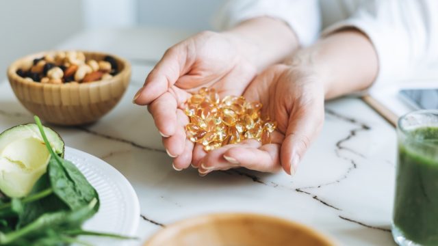 Close up of a woman's hands holding supplements surrounded by greens, nuts, and a green smoothie