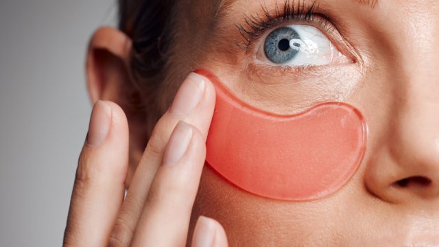 Closeup of a woman applying a peach under eye patch against a gray background