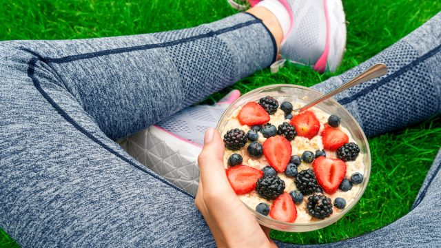View of woman's legs in gray workout leggings while sitting in the grass holding a bowl of oatmeal and berries
