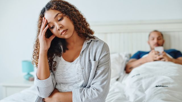 An upset-looking woman sitting on the edge of the bed with her husband in the background playing on his phone