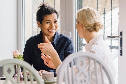 Two smiling women are sitting at a table in a cafe, socialising over tea.