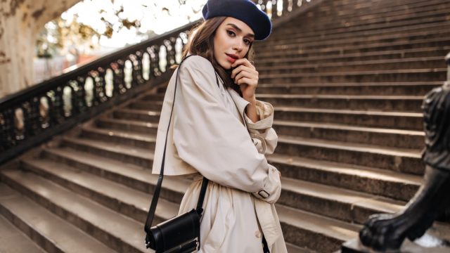 Lovely young Parisienne with brunette hair in stylish beret, beige trench coat and black bag, standing on old stairs and sensitively posing outdoors.