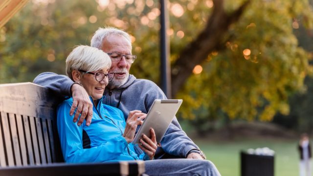 Smiling senior active couple sitting on the bench looking at tablet computer. Using modern technology by elderly.