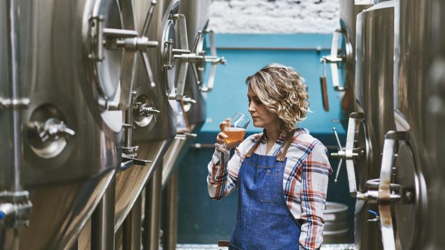 A female brewer sampling her beer while standing next to fermenters in a brewery