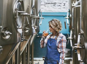 A female brewer sampling her beer while standing next to fermenters in a brewery