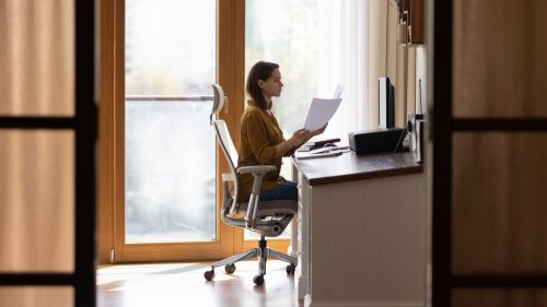 Serious home distance employee doing paperwork at table, reviewing financial paper reports at desktop computer, working at comfortable safe workplace with ergonomic office chair. Full length shot