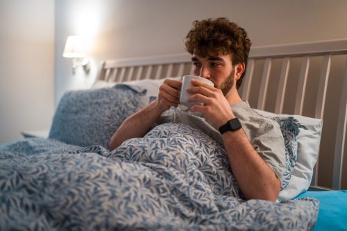 male in casual nightwear, holding a cup, lying in bed unable to sleep, depicting insomnia indoors at night with a concerned expression.