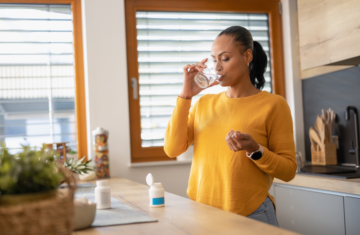 woman in a yellow sweater taking medicine or vitamin with water in her kitchen