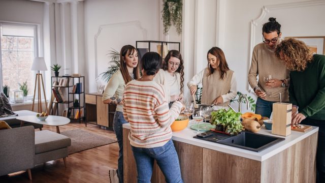 Group of people, diverse male and female friends preparing a meal and drinking wine together in domestic kitchen,