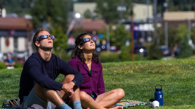 Man and woman sitting on grass while watching a solar eclipse with protective glasses