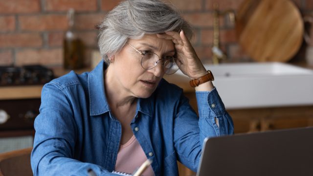 A senior woman sitting at her laptop in a kitchen with a distressed look on her face