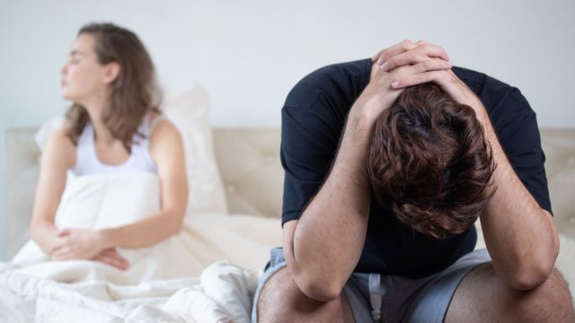 man sitting on bed with his head in hands, woman sitting in bed looking away