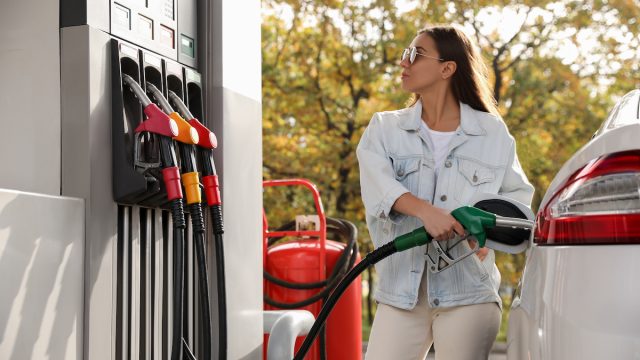 Young woman pumping gas into her white car, looking at the screen at the gas station pump