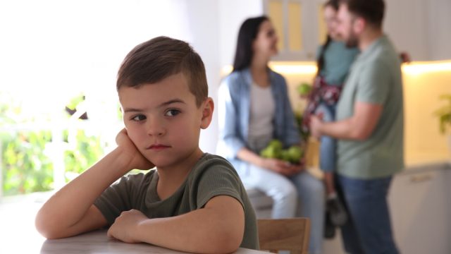 Unhappy little boy feeling jealous while parents spending time with his sister at home