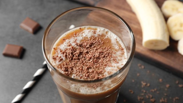 Glass with chocolate protein shake on table, with banana in the background
