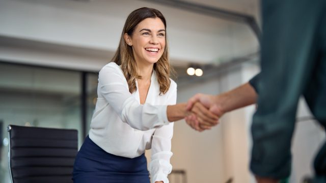 Happy mid aged business woman manager handshaking greeting client in office. Smiling female executive making successful deal with partner shaking hand at work standing at meeting table