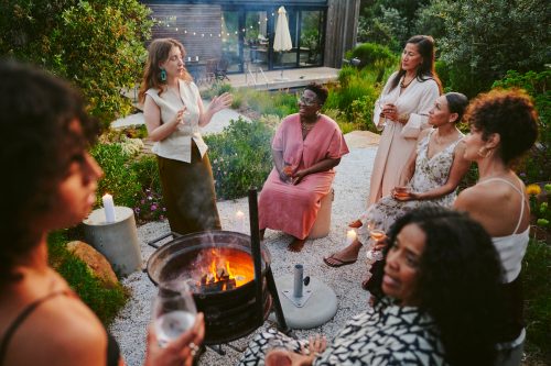 group of smiling women having drinks and talking around a fire pit during an early evening party in the lush garden of a home at dusk
