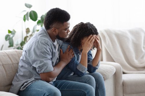 Dependent Relationship Concept. Young Guy Soothing His Depressed Girlfriend At Home, Embracing Her On Couch In Living Room