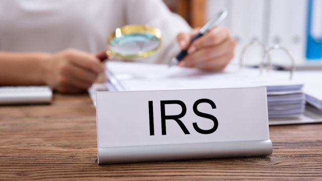 Close-up Of Nameplate With IRS Title Kept On Desk In Front Of Businesswoman Working