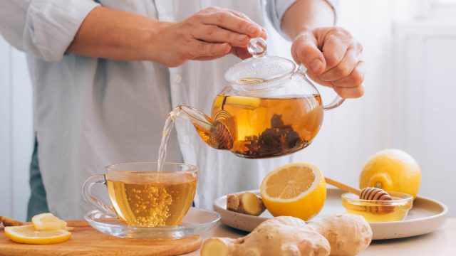 Cropped shot of a person pouring lemon-ginger tea from a glass teapot into a glass mug on a table surrounded by lemons and ginger