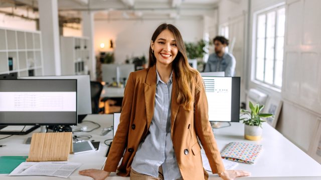 young woman in business-casual outfit leaning on a desk in an office
