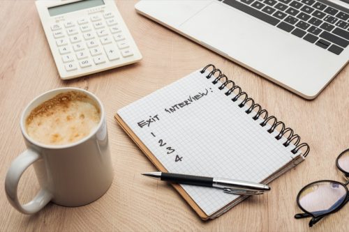 Notebook with exit interview lettering and numbers on wooden table with laptop, stationery, glasses and cup of coffee